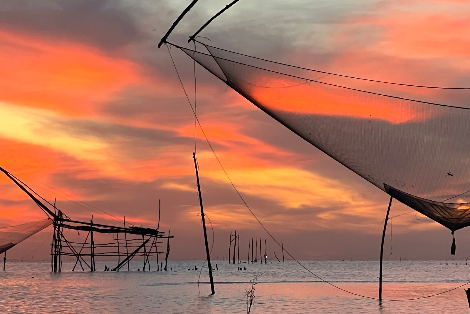 Traditional giant lift nets silhouetted against an orange sunset over a lake in southern Thailand